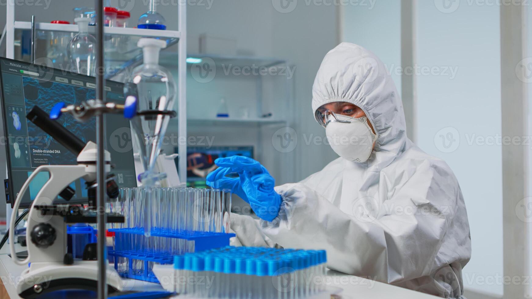 Lab technician in coverall examining samples of liquid typing on computer results sitting in equipped laboratory. Scientist working with various bacteria tissue pharmaceutical research for antibiotics photo