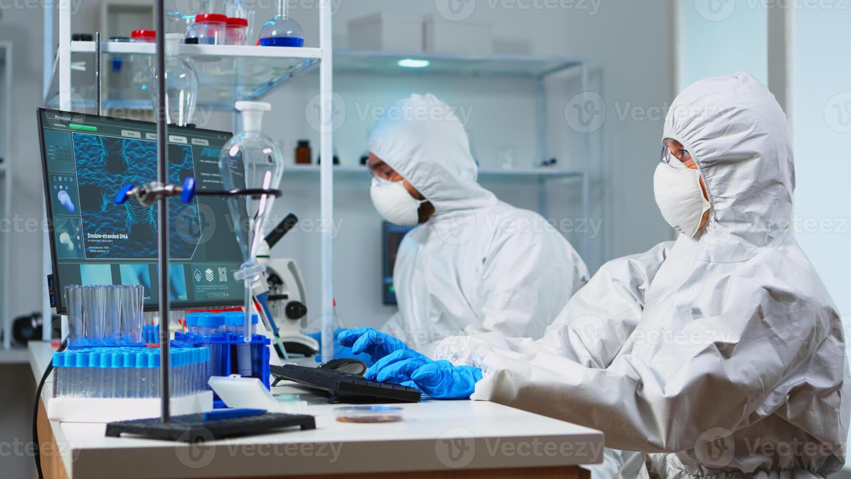 Team of biologists in coverall working with microscope and typing on computer sitting in laboratory. Scientists examining vaccine evolution using high tech researching diagnosis against covid19 virus photo