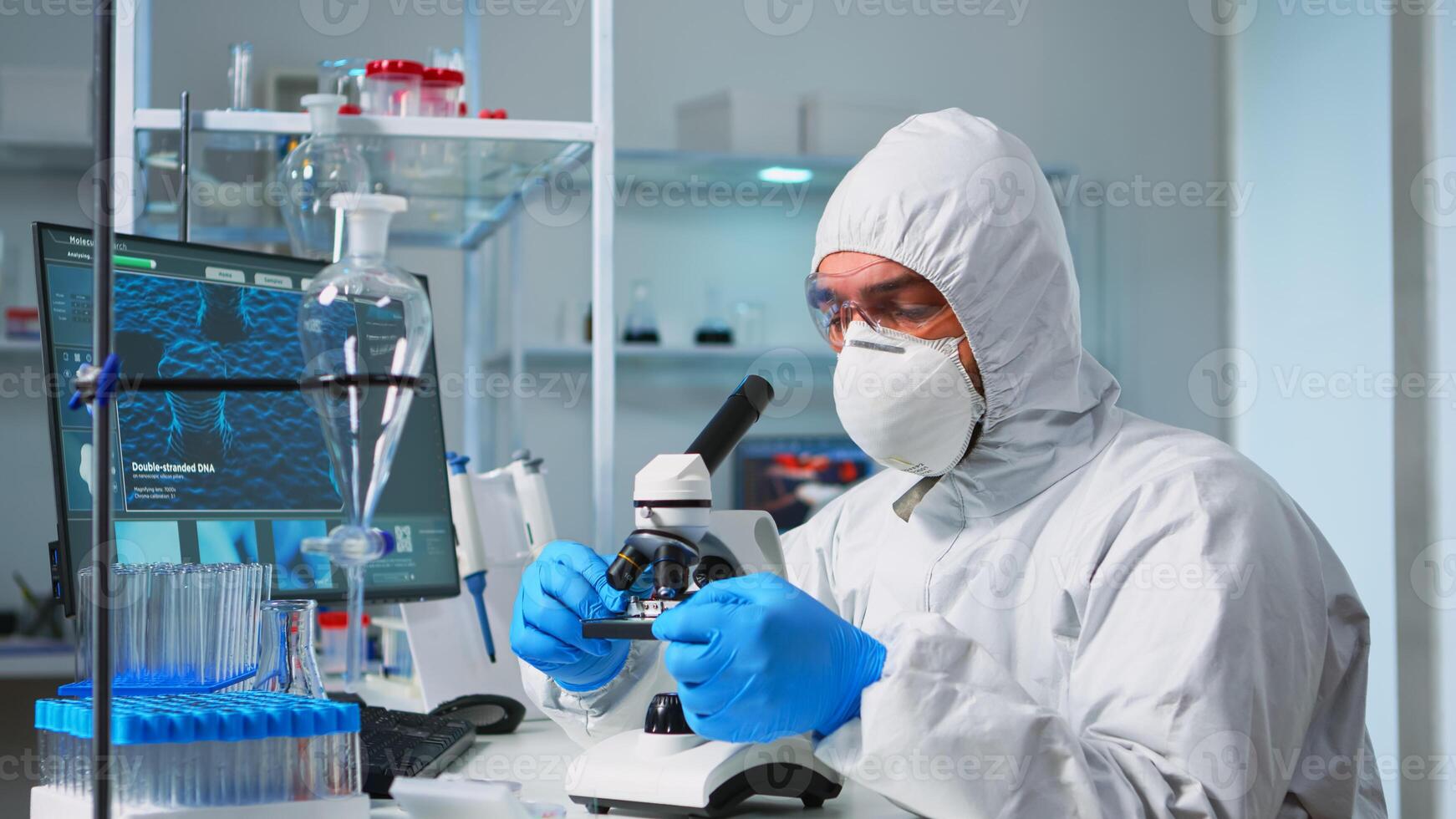 Scientist in ppe suit placing a slide on specimen stage of a laboratory microscope making adjustments. Chemist in coverall working with various bacteria, tissue blood samples for antibiotics research photo
