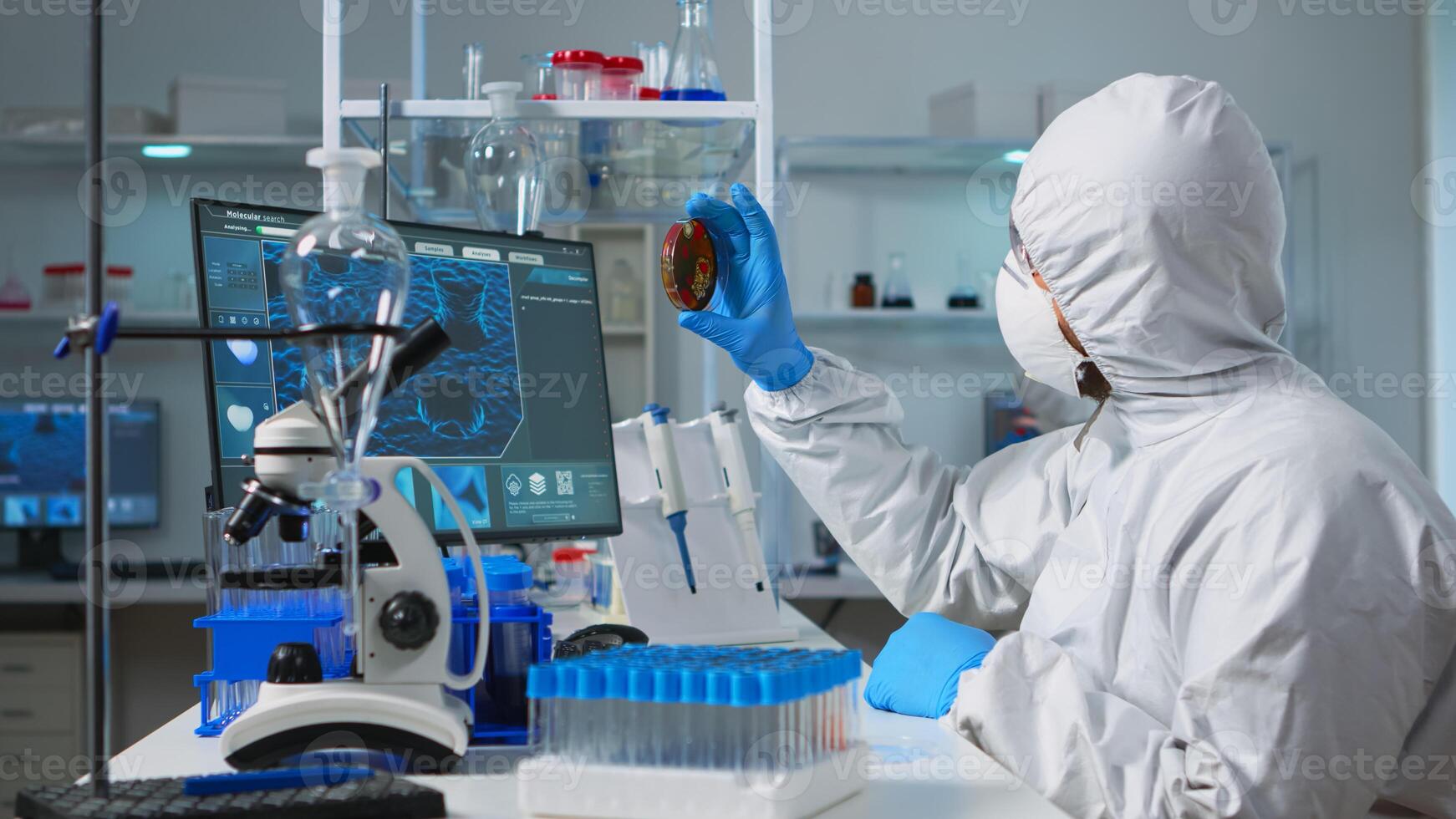 Chemist with coverall analysing bacteria sample from petri dish typing results. Team of doctors examining vaccine evolution with high tech, chemistry tools for research in development of covid19 virus photo