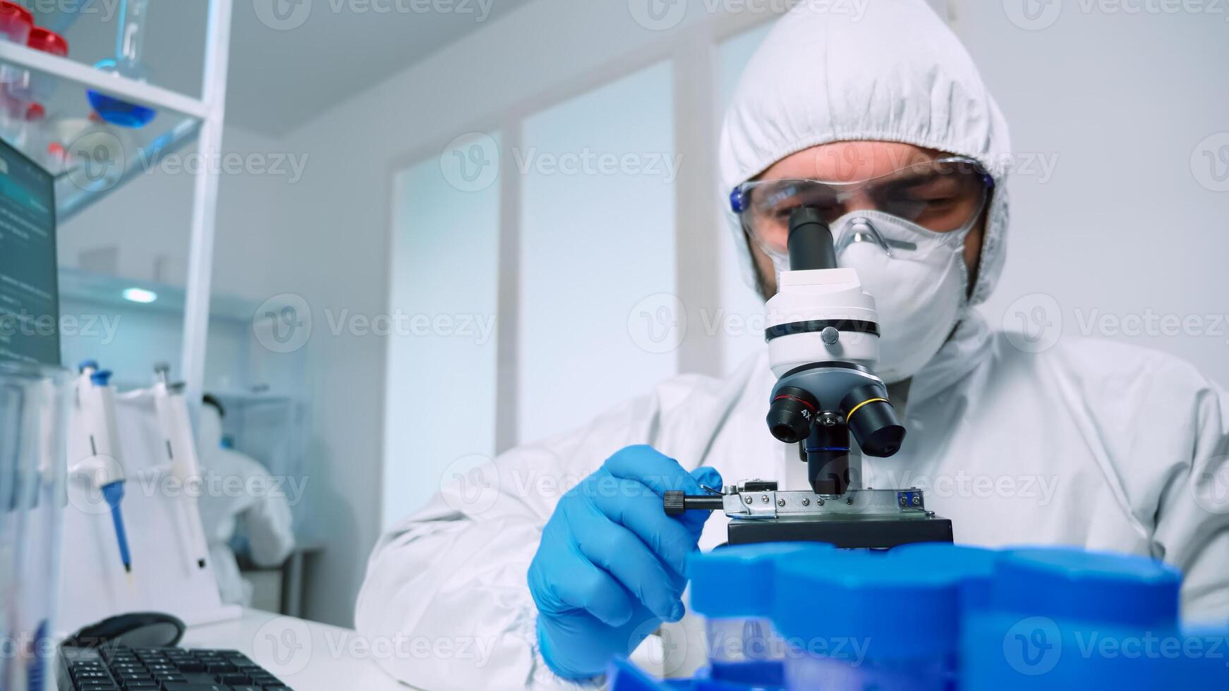 Laboratory scientist in ppe suit conducting experiment looking through a microscope in modern lab. Man working with various bacteria tissue, blood samples, concept of medical research for antibiotics photo