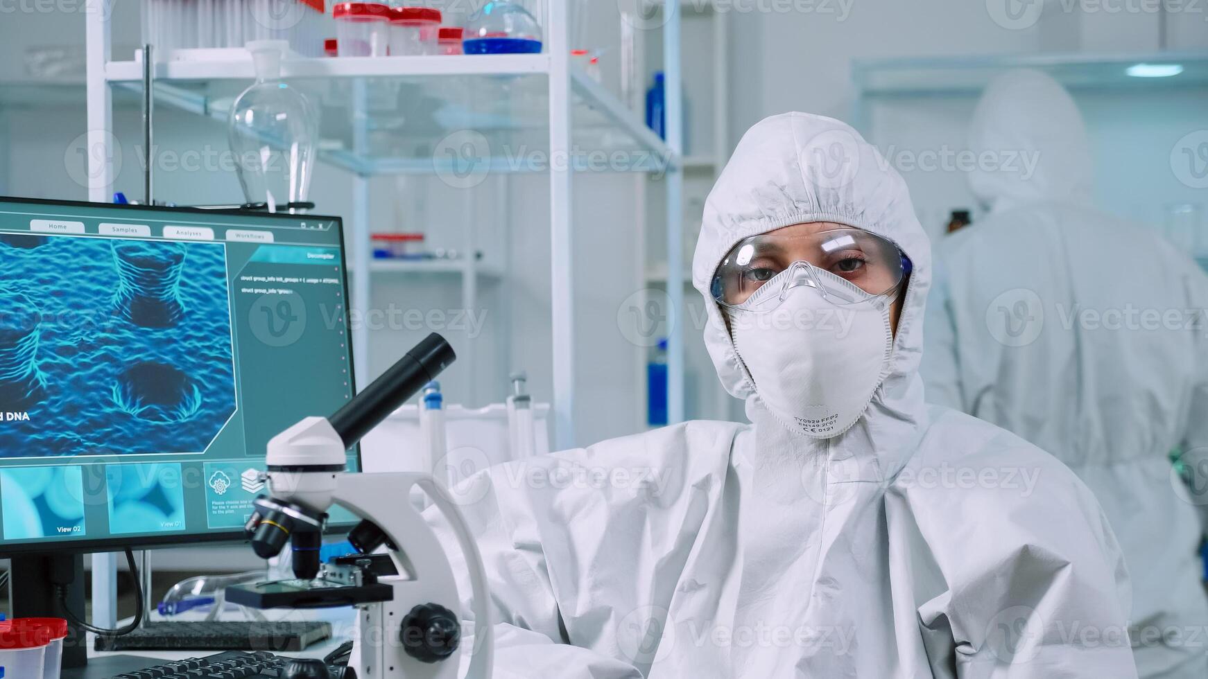 Tired laboratory doctor with overall suit looking at camera in equipped lab. Team of scientists examining virus evolution using high tech, chemistry tools for scientific research, vaccine development photo