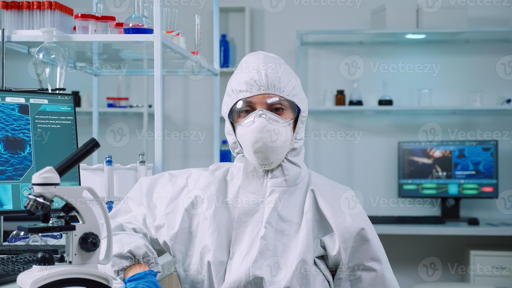Tired microbiologist in coverall sitting in laboratory looking at camera. Team of scientits examining virus evolution using high tech and chemistry tools for scientific research, vaccine development. photo