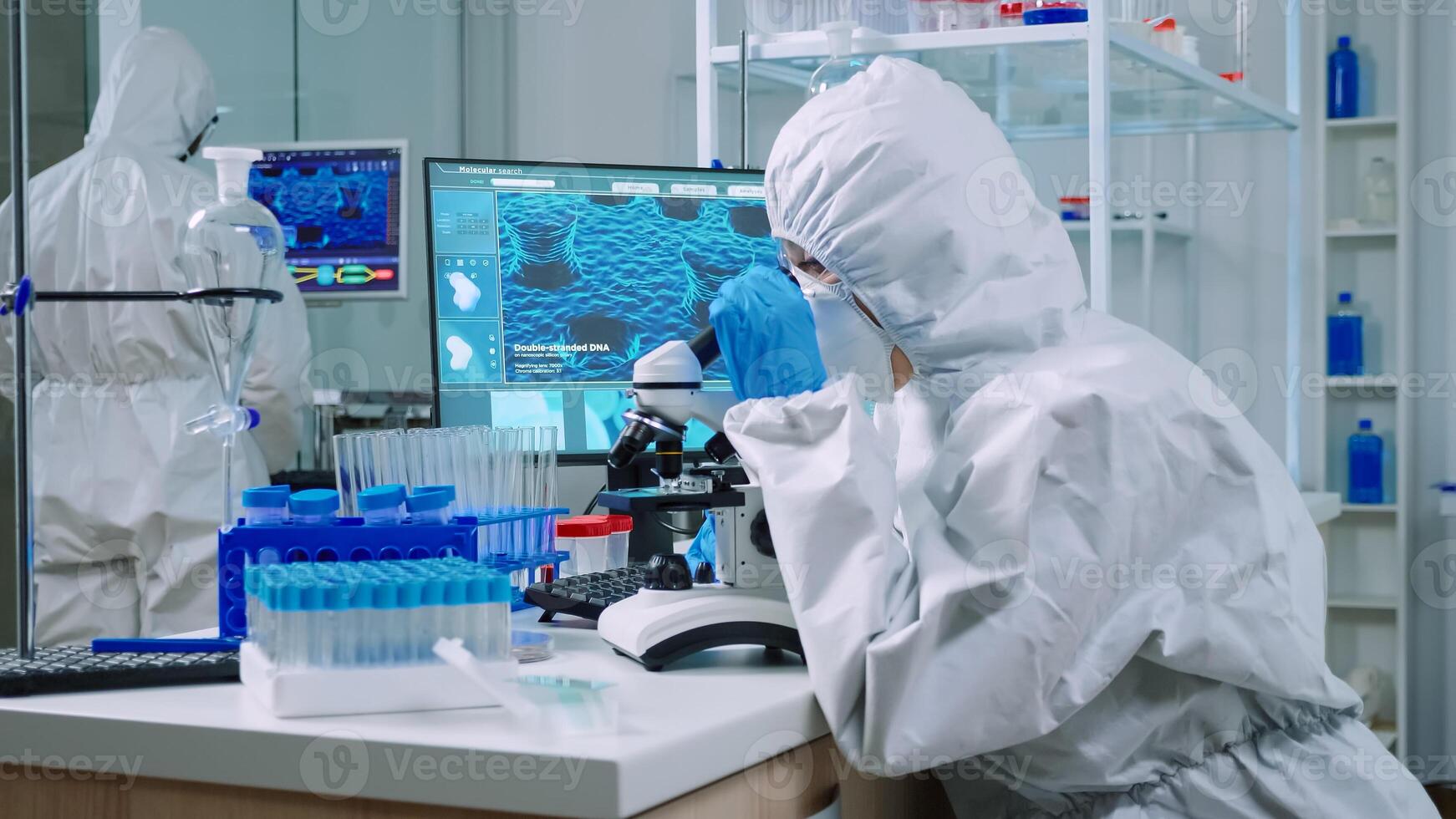 Chemist in protection suit typing on pc and analyzing virus sample using microscope in scientific lab. Team of doctors examining vaccine evolution using high tech researching diagnosis against covid19 photo