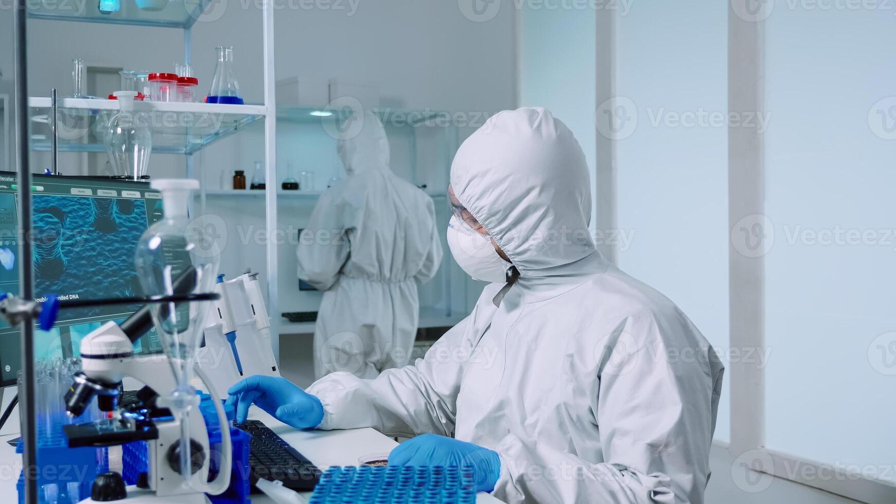 Scientist in ppe suit holding petri dish while studying virus samples in biotechnology lab typing on pc. Doctors examining vaccine evolution using high tech researching diagnosis against covid19 virus photo