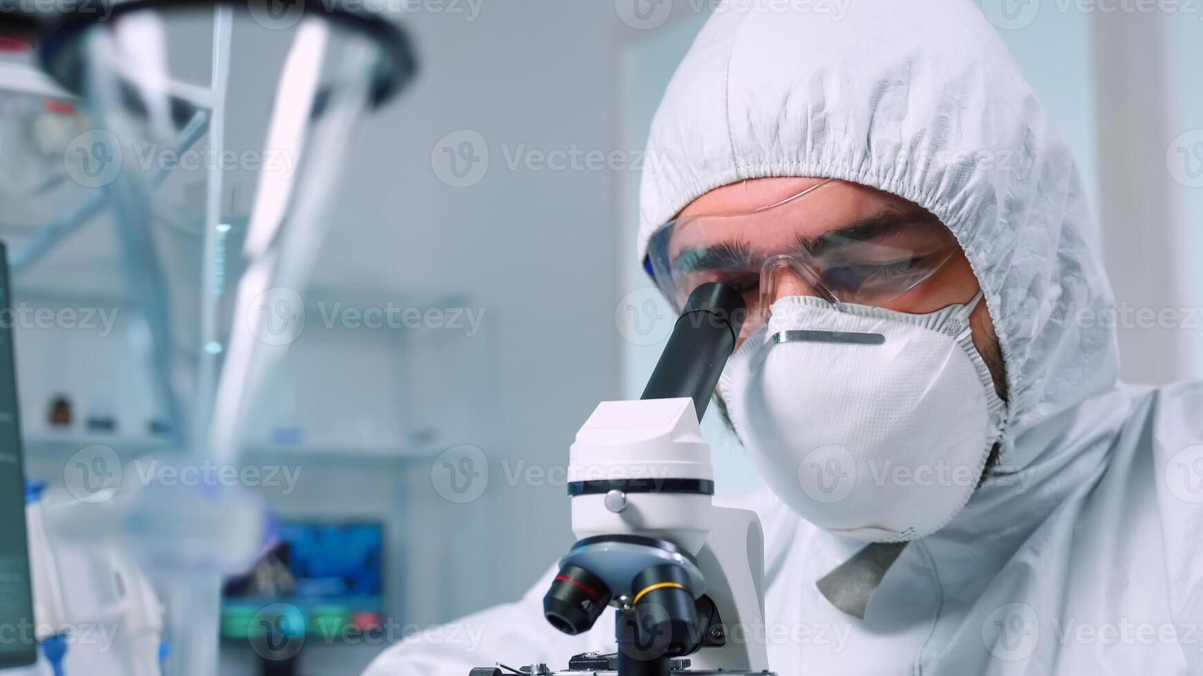 Biochemist in ppe suit looking through a microscope during experiment in sterile lab. Chemist researcher examining virus evolution using high tech for vaccine scientific research against covid19 photo