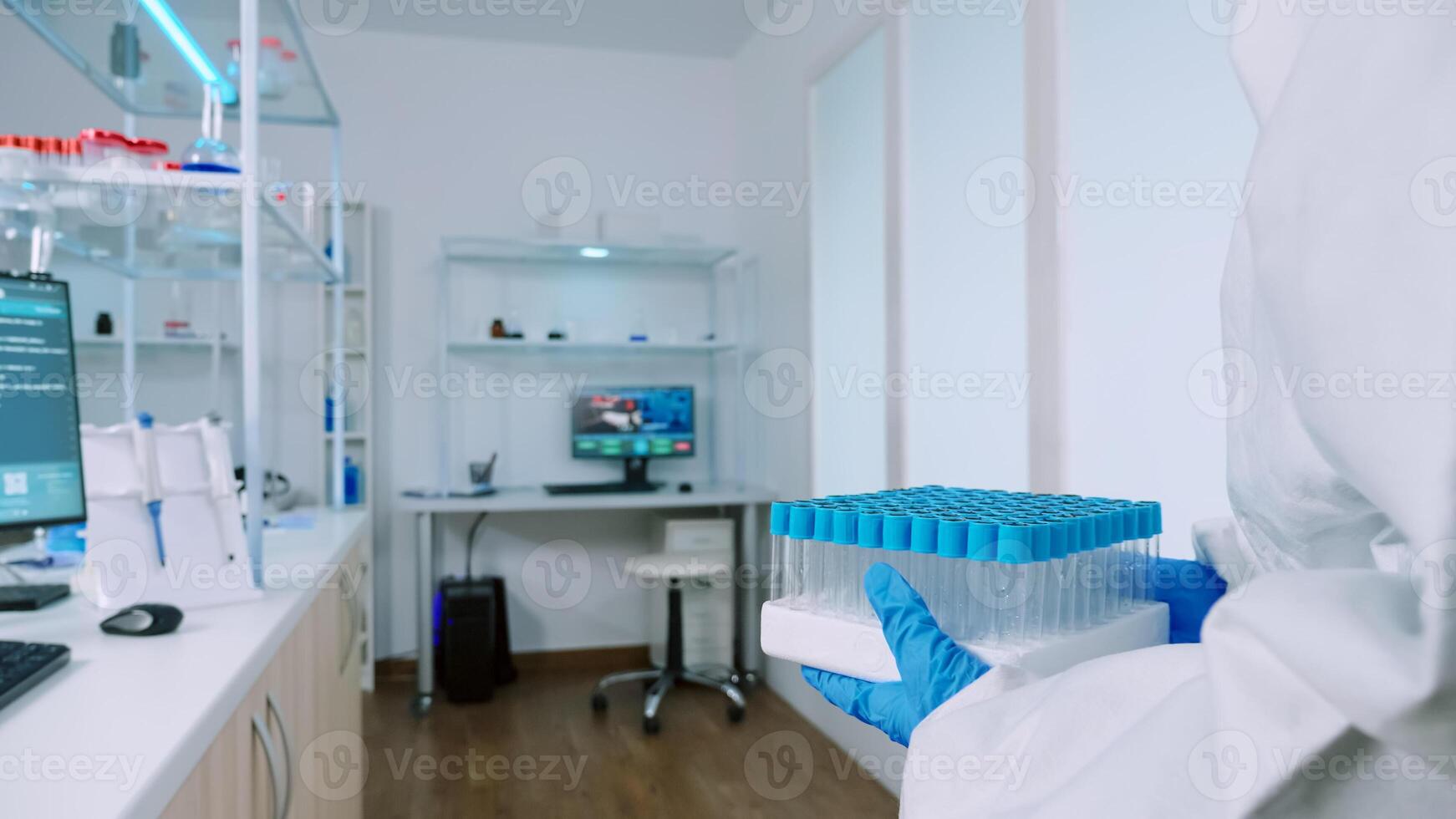 Chemist in protection suit bringing test tubes in steril laboratory and typing data on computer. Doctors examining virus evolution using high tech for research of treatment development against covid19 photo