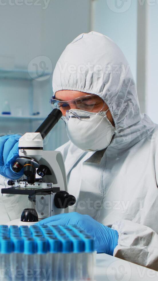 Medical research scientist in ppe suit conducting vaccine development using digital microscope in a biological applied science laboratory. Caucasian lab engineer in coverall working on medical treatment photo