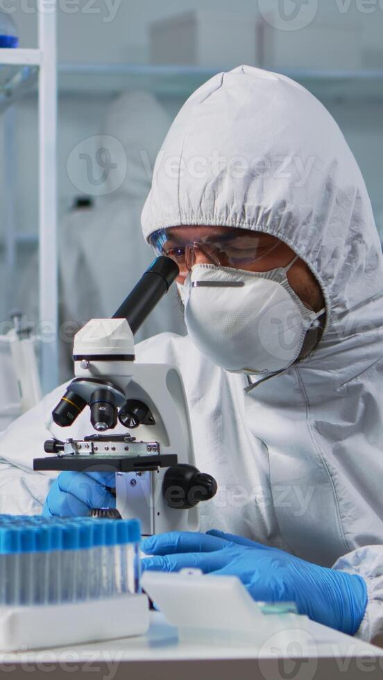 Close up of scientist looking through a microscope wearing ppe suit in equipped lab. Team examining virus evolution using high tech for scientific research of vaccine development against covid19. photo