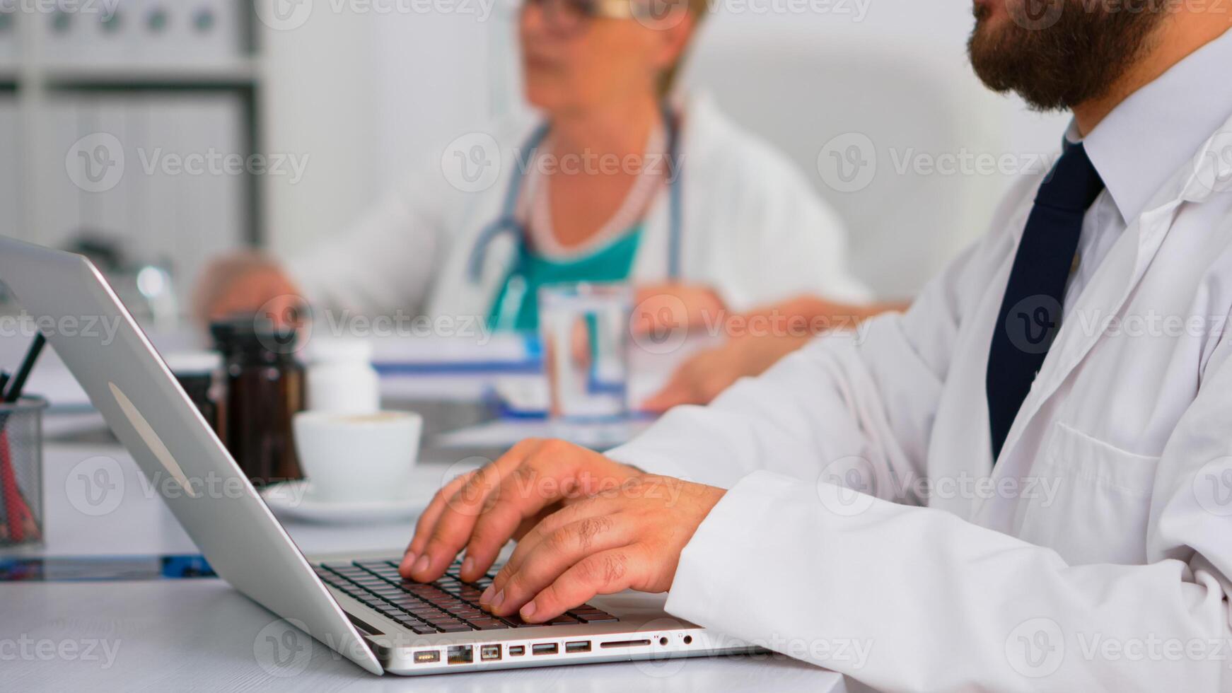 Close up of doctor using laptop, writing treatment informations while coworkers discussing in background during medical conference sitting at desk in hospital office. Team of doctors brainstorming photo