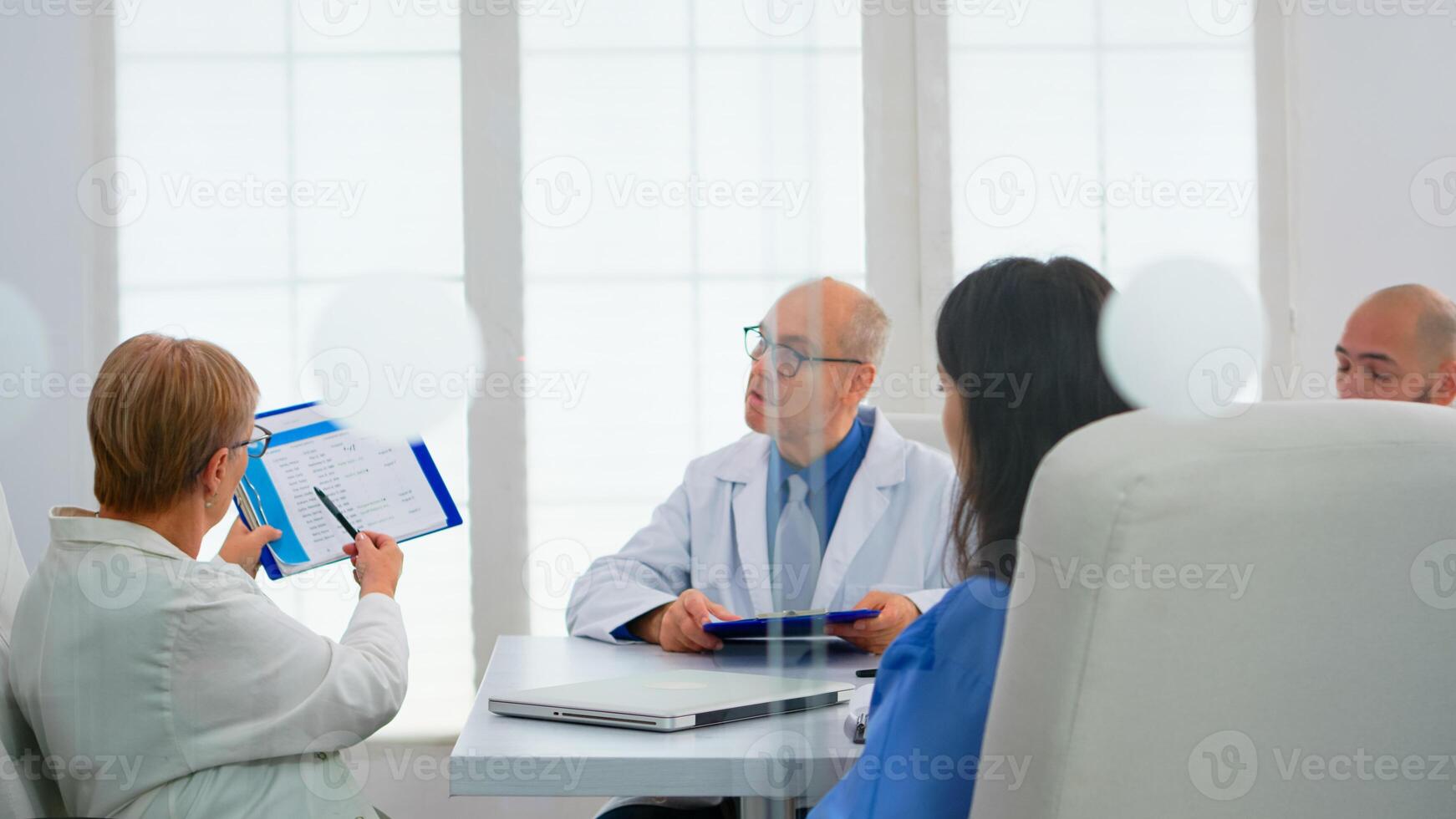 Elderly doctor having medical conference in hospital meeting room discussing medical problems pointing on clipboard presenting list of sick patients. Team of doctors presenting symptoms of disease photo