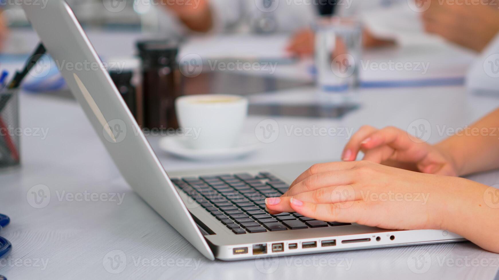 Close up of assistant using laptop, writing meeting informations while doctors discussing in background during medical conference sitting at desk in hospital office. Profesional team brainstorming photo