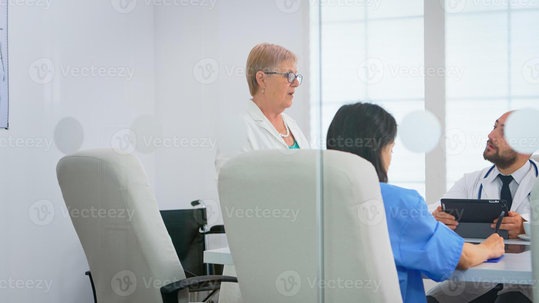 Medical team sitting and discussing at table in hospital meeting office using tablet and clipboard with documents of patients. Group of physicians talking about symptoms of disease in clinic office photo