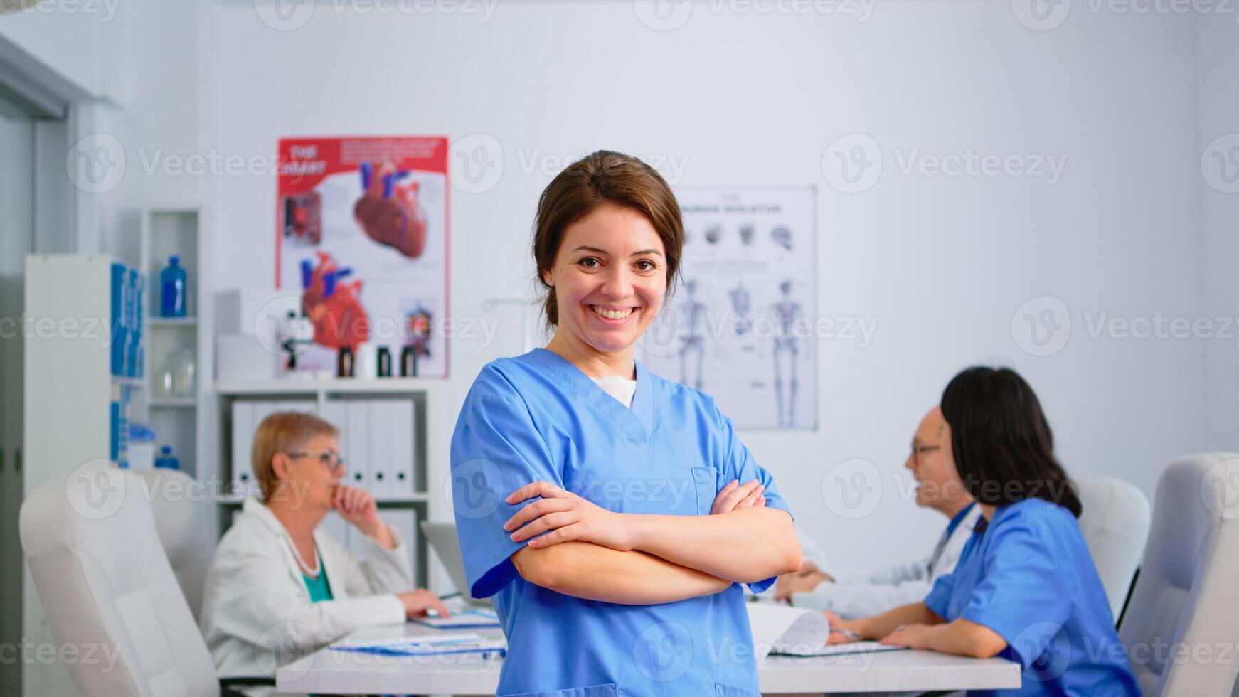 Portrait of young nurse standing in front of camera smiling in medical conference meeting office. Team of specialist doctors working in background talking about symptoms of disease in clinic room photo