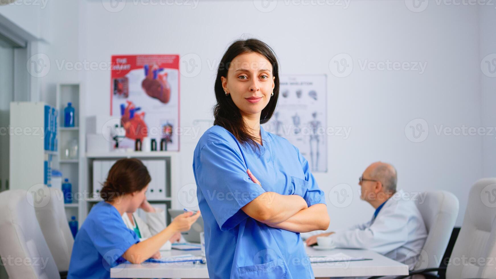 Nurse standing in front of camera smiling in medical conference meeting office. Team of specialist doctors working in background talking about symptoms of disease in clinic room photo