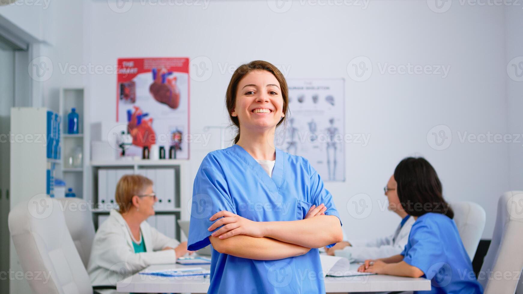 Nurse looking at camera smiling standing in medical conference meeting office. Team of specialist doctors working in background talking about symptoms of disease in clinic room photo