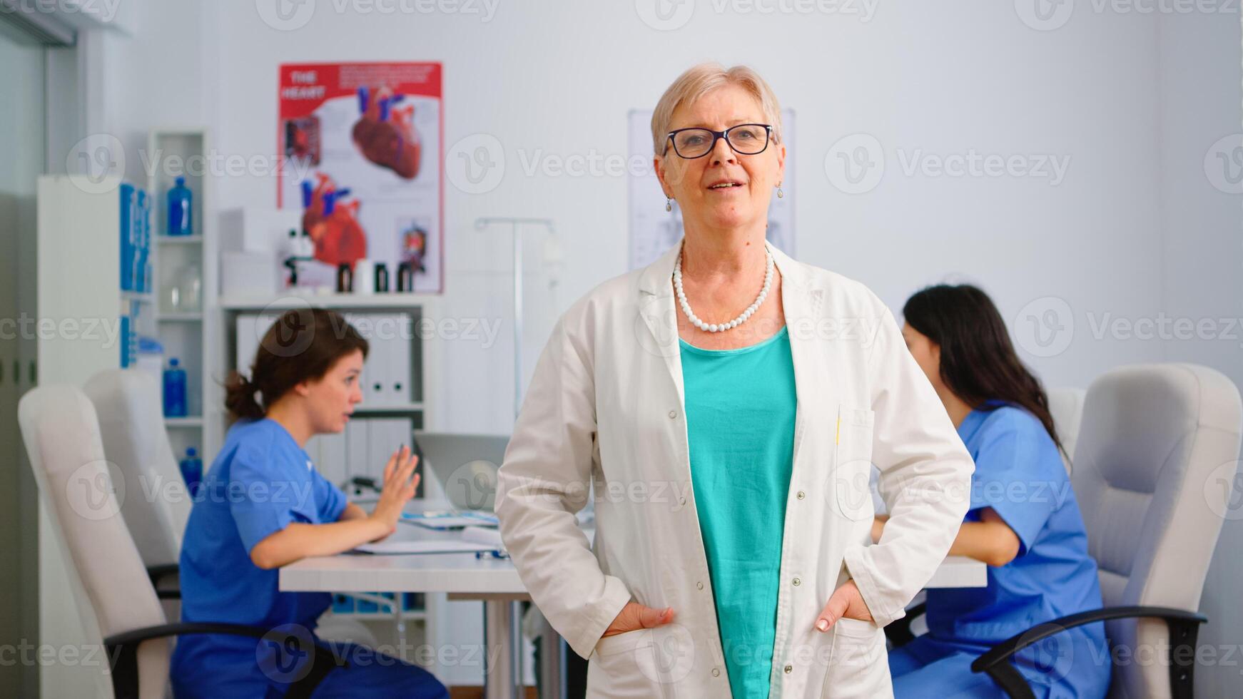 Portrait of elderly doctor standing in front of camera smiling in medical conference meeting office. Team of specialist doctors working in background talking about symptoms of disease in clinic room photo