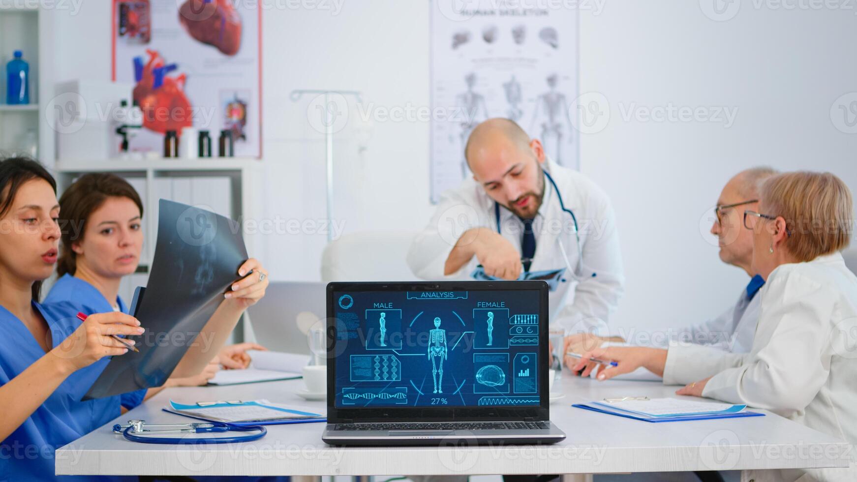Team of doctors with diverse ages brainstorming sitting on desk in hospital meeting office placing laptop with human body image in front of camera. Nurse talking about symptoms of disease in clinic. photo