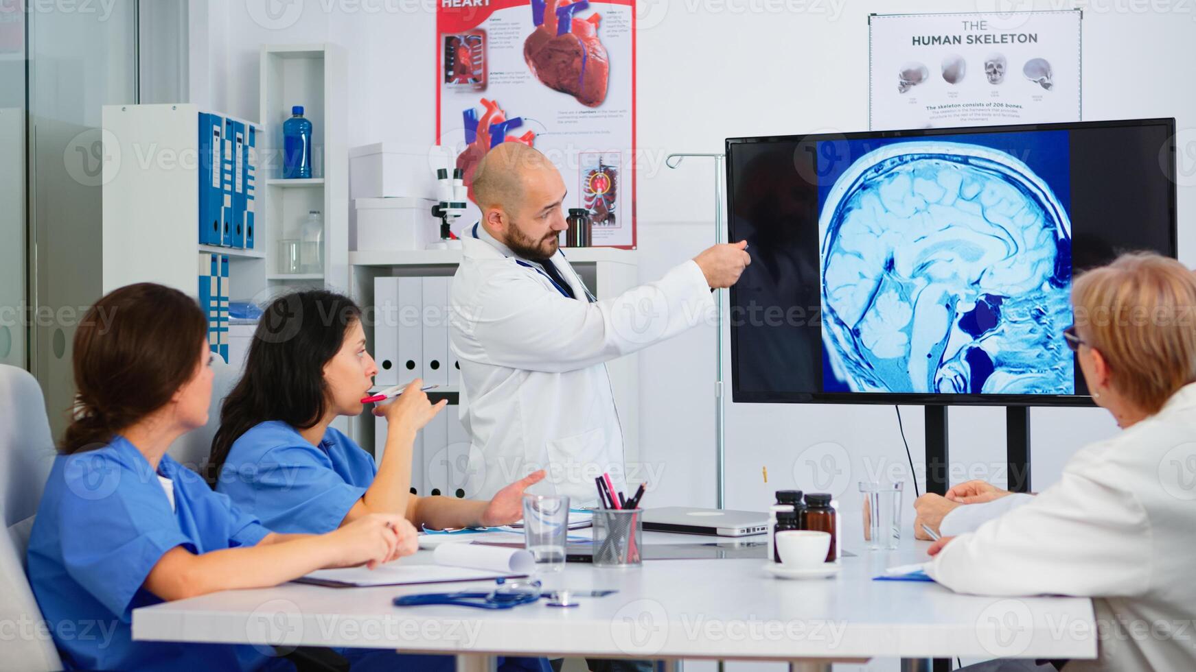 Experienced man doctor analysing the result of digital x-ray image together with cvalified colleagues in meeting room, pointing on monitor. Doctors discussing diagnosis about treatment of patients photo