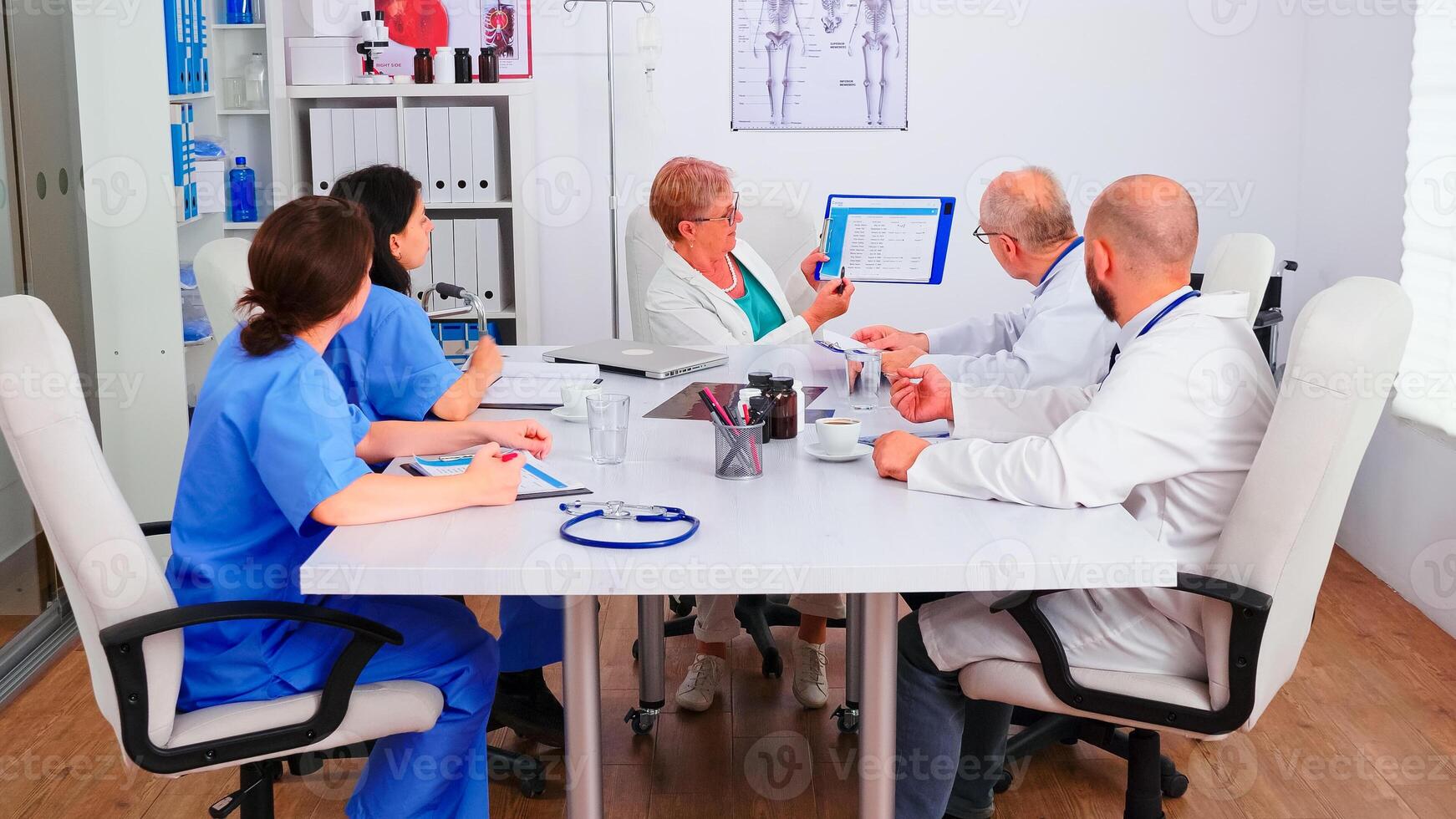 Experienced doctors in hospital meeting room working together during health seminar, senior medic pointing on clipboard. Expert therapist talking with colleagues about disease, medicine professional photo