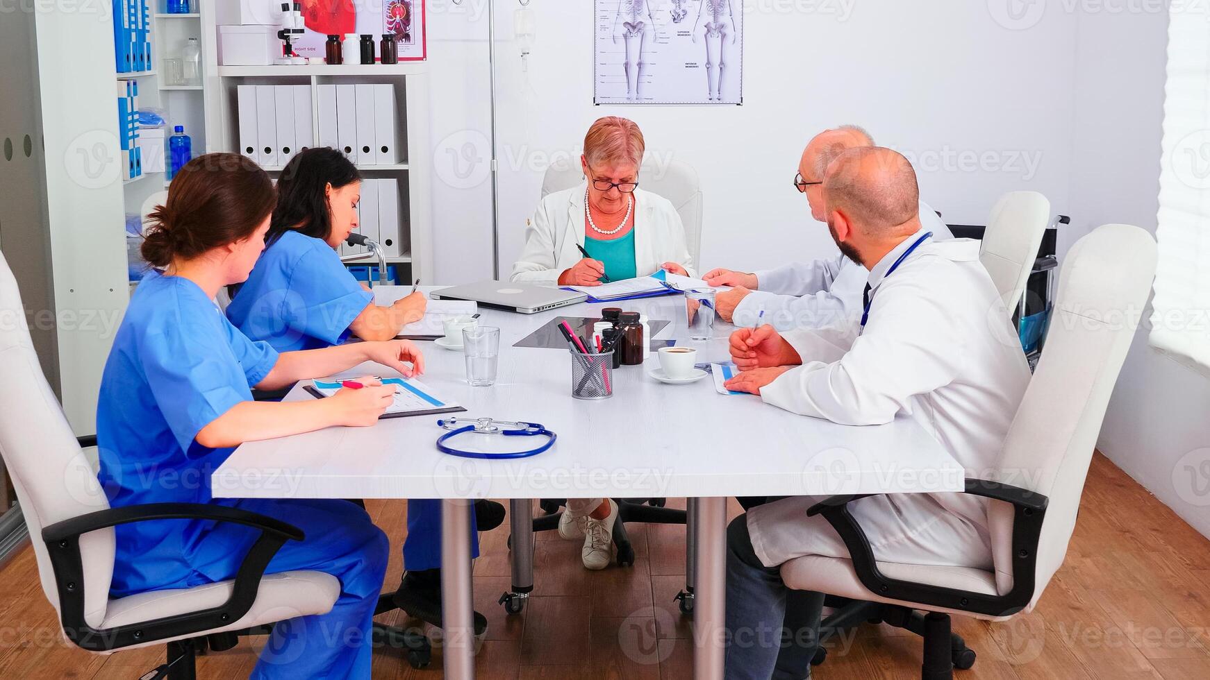 Medical nurse writing on clipboard during healthcare seminar in hospital meeting office about client's list. Clinic expert therapist talking with colleagues about disease, medicine professional. photo
