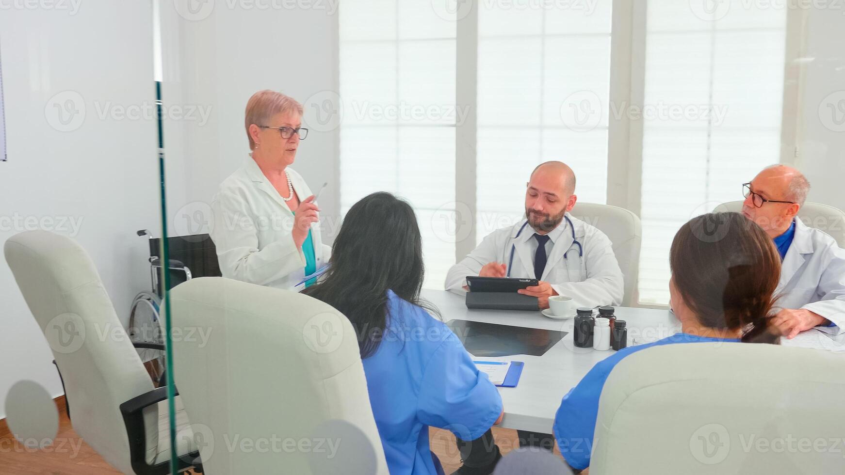 Expert elderly woman doctor having a discussion with medical staff in hospital conference room. Clinic expert therapist talking with colleagues about disease, medicine professional photo