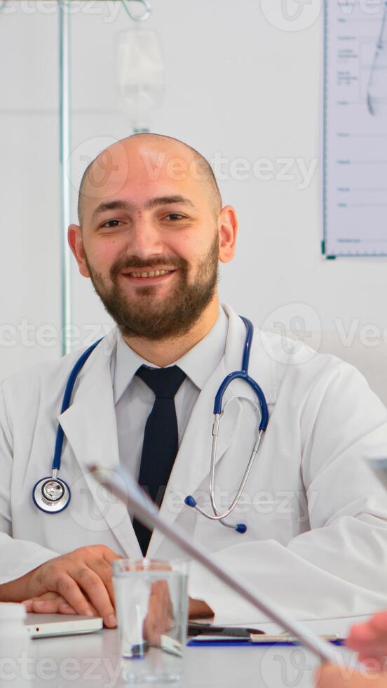 Doctor in lab coat and stethoscope over neck typing on laptop at desk looking at camera and smiling in busy workday in medical office. Specialist writing on notepad during brainstorming in meeting room photo