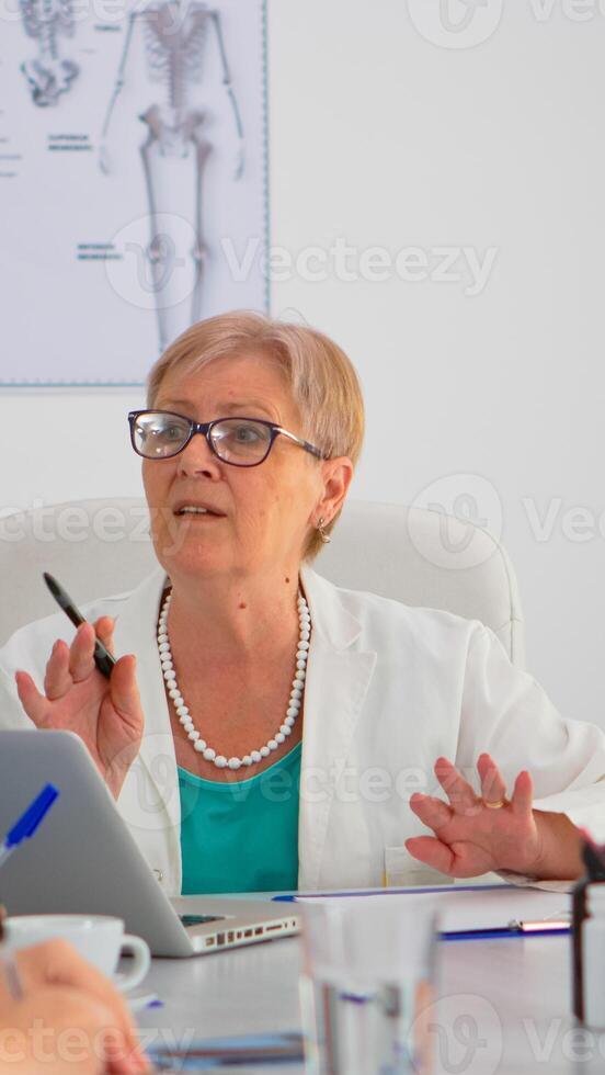 Senior surgeon explaining treatment to coworkers presenting plan during medical conference sitting at meeting desk in modern clinic. Profesional team taking notes while specialist doctor discussing. photo