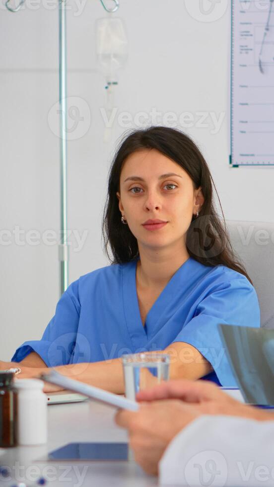 Portrait of nurse typing on laptop during brainstorming and looking at camera smiling sitting on desk in hospital office, conference room. Specialist in lab coat writing on notepad in medical clinic. photo
