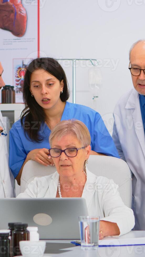 Medical team discussing diagnosis about patients treatment issues record in workplace using laptop. Group of doctors, teamwork people having health brainstorming conference in hospital meeting room. photo