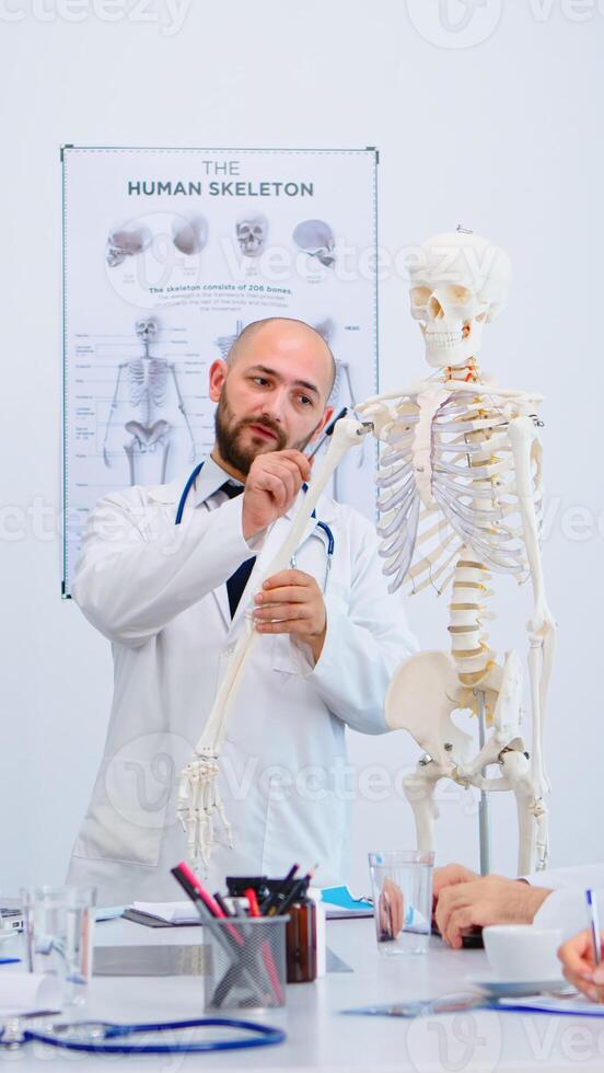 Medical doctor man pointing on cervical spine of human skeleton anatomical model, explaining to coworkers medical procedures in hospital meeting room. Physicians discussing about symptoms of disease photo
