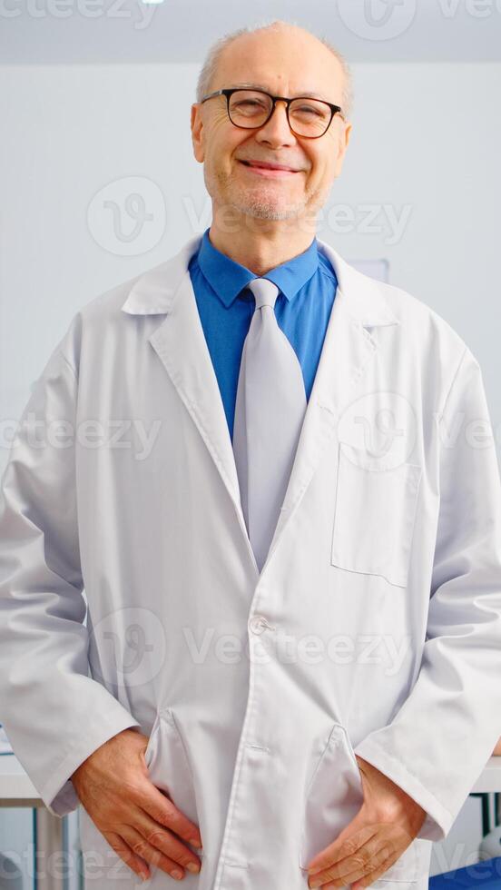 Portrait of elderly man surgeon standing in front of camera smiling in medical conference meeting office. Team of doctors working in background talking about symptoms of disease in clinic room photo