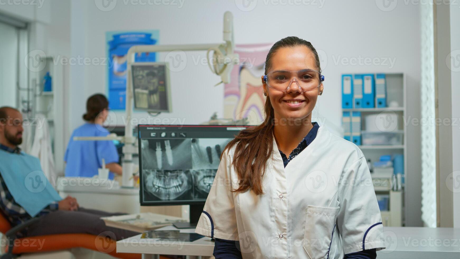 Stomatologist doctor smiling at camera in dental office while assistant preparing man for stomatological surgery in background. Woman dentist looking on webcam sitting on chair in stomatological room photo
