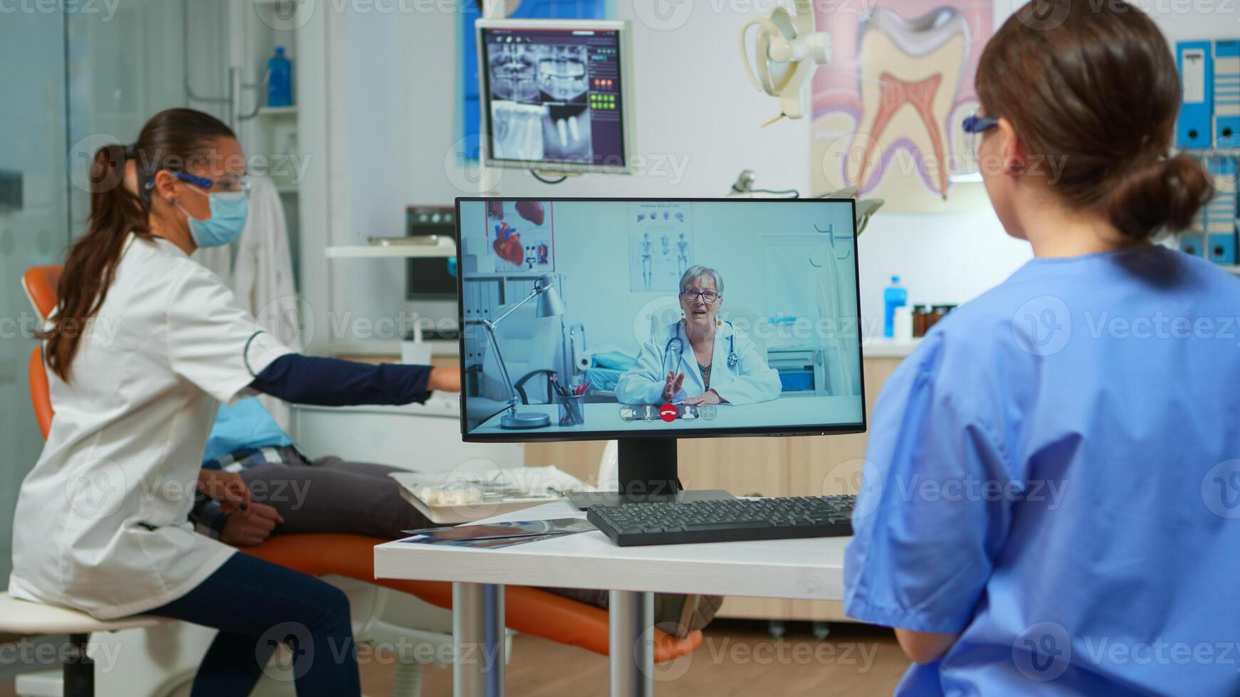 Assistent having call with expert stomatologic dotor using computer while doctor is working with patient in background. Nurse listening dentist on webcam sitting on chair in stomatological office photo