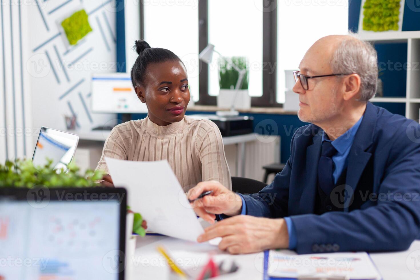 African employee discussing with senior executive looking financial charts in startup company conference board room. Multiethnic businesswoman and mature entrepreneur brainstorming. photo