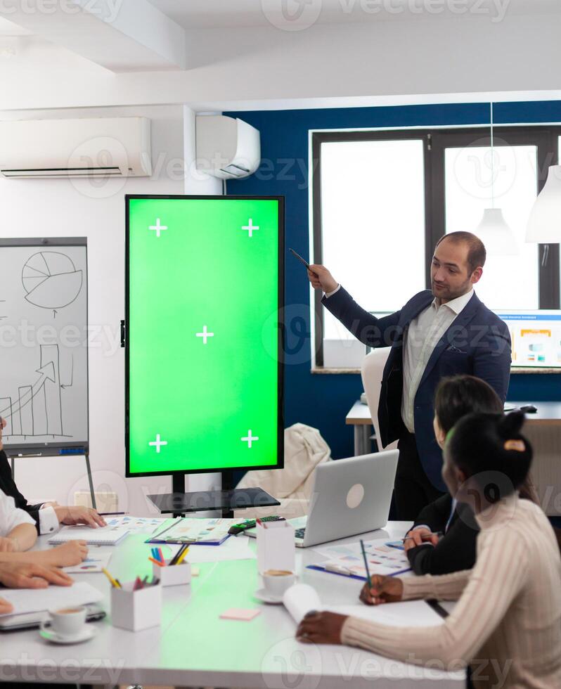 Leader of company presenting financial plan using mockup display in front of diverse team brainstorming. Manager explainig project strategy on green screen monitor with chroma key desktop in boardroom photo