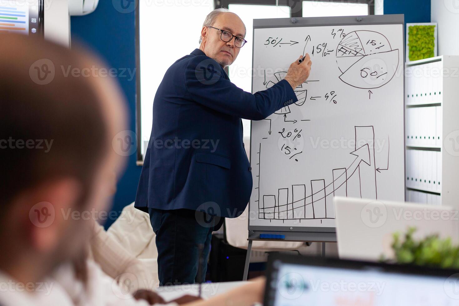 Elderly businessman giving presentation to multiethnic team of colleagues using flip chart. Serious speaker boss executive, business trainer explaining development strategy to motivated mixed race employees. photo