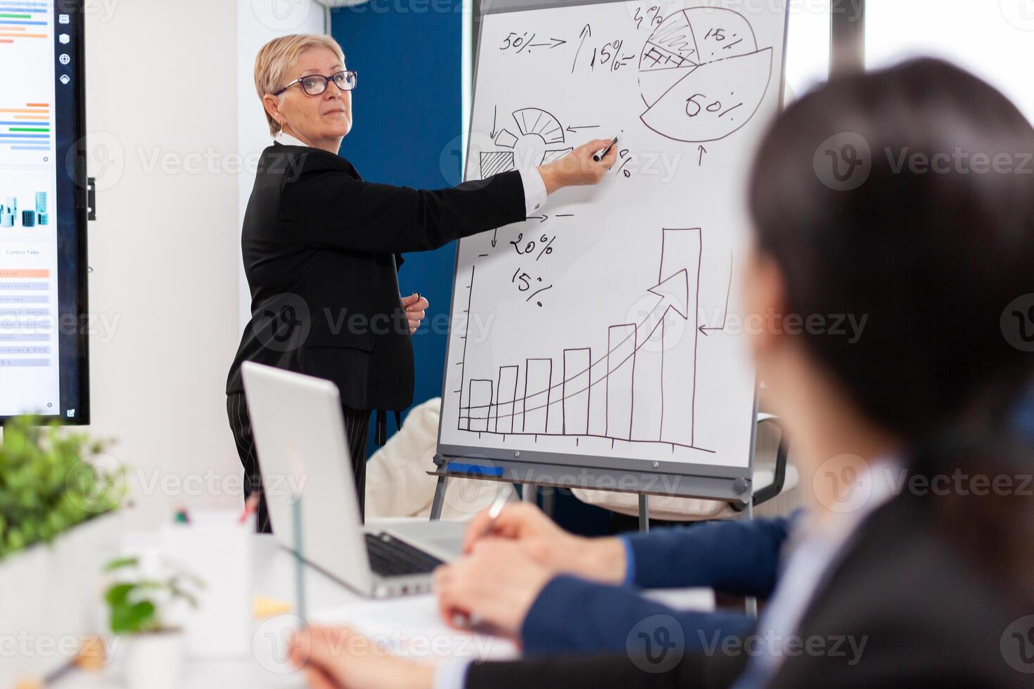 maduro mujer de negocios escritura en blanco junta, presentación ventas evolución respondiendo pregunta. grave altavoz jefe ejecutivo, negocio entrenador explicando desarrollo estrategia a motivado mezclado carrera empleados. foto