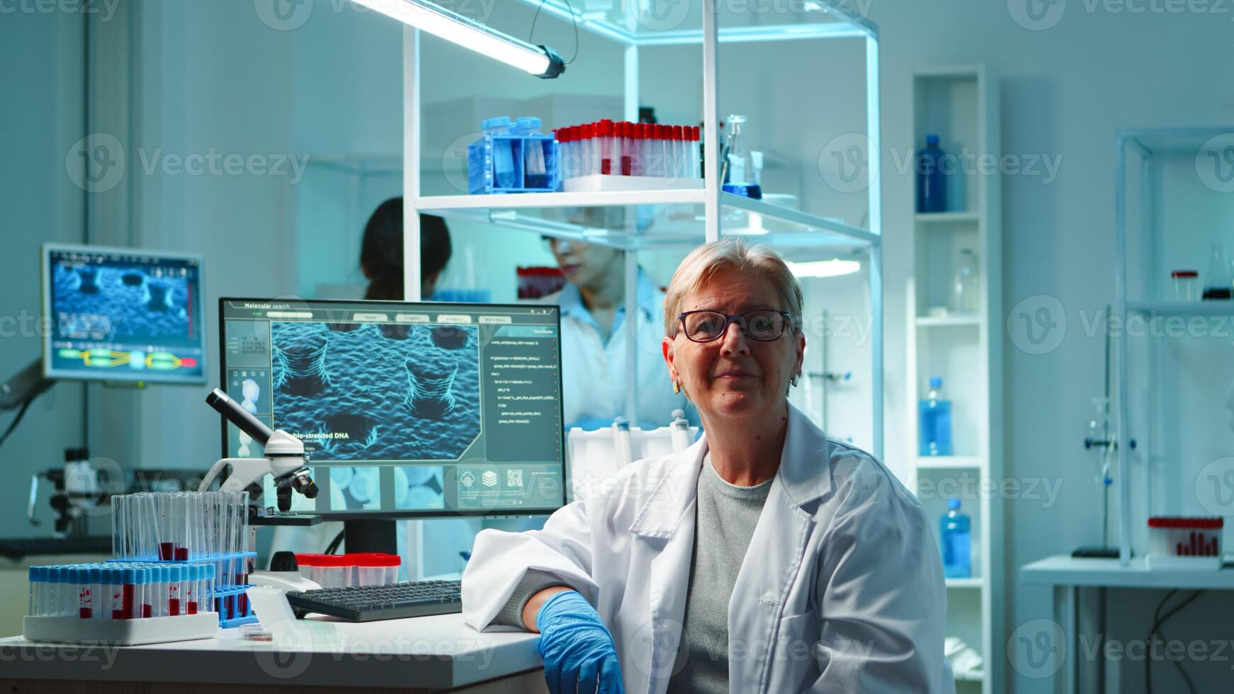 Portrait of senior scientist woman looking exhausted at camera sitting in equipped lab late at night. Team examining virus evolution using high tech for scientific research, vaccine development photo