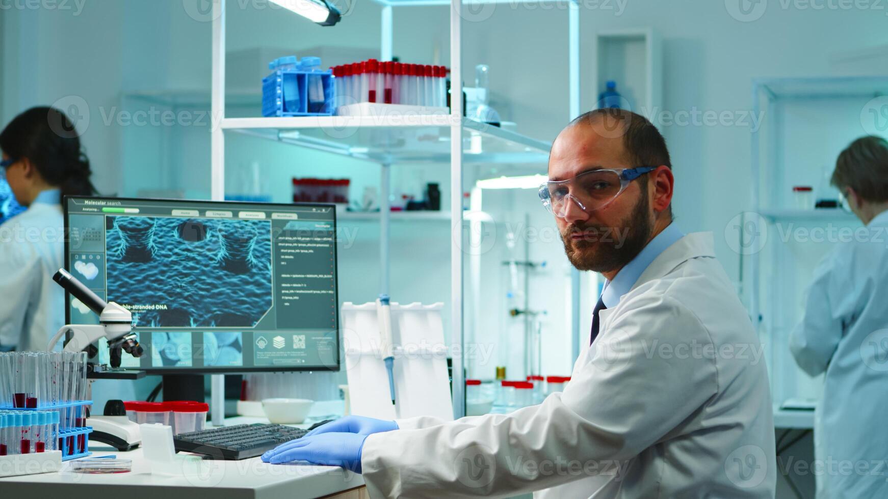 Worn out doctor typing on computer and looking exhausted at camera in equipped lab late at night. Medical chemists examining virus evolution using high tech for scientific research, vaccine development. photo
