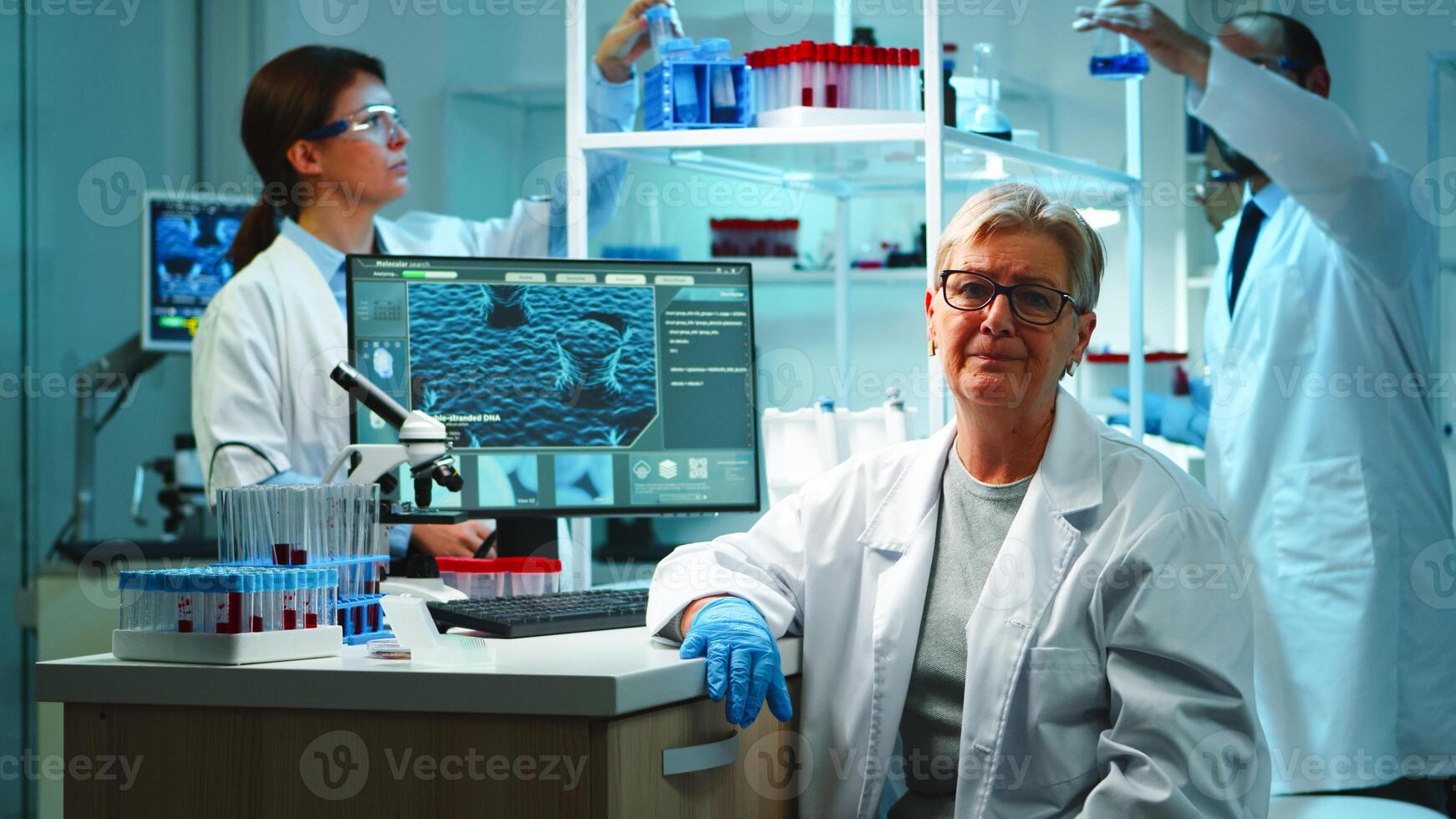 Portrait of old chemist woman with fatigued face in front of camera in equipped lab late at night. Team examining virus evolution using high tech and chemistry tools for research, vaccine development photo