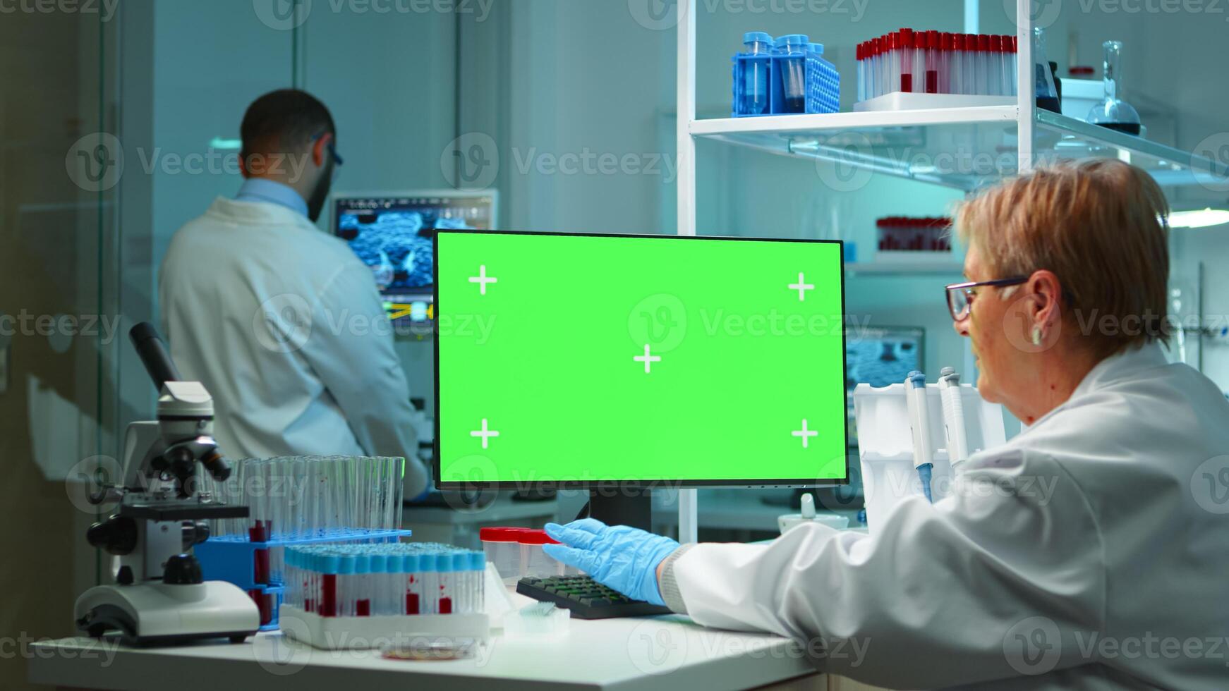 Senior doctor working at computer with green screen in modern equipped lab late at night. Team of microbiologists doing vaccine research writing on device with chroma key, isolated, mockup display. photo