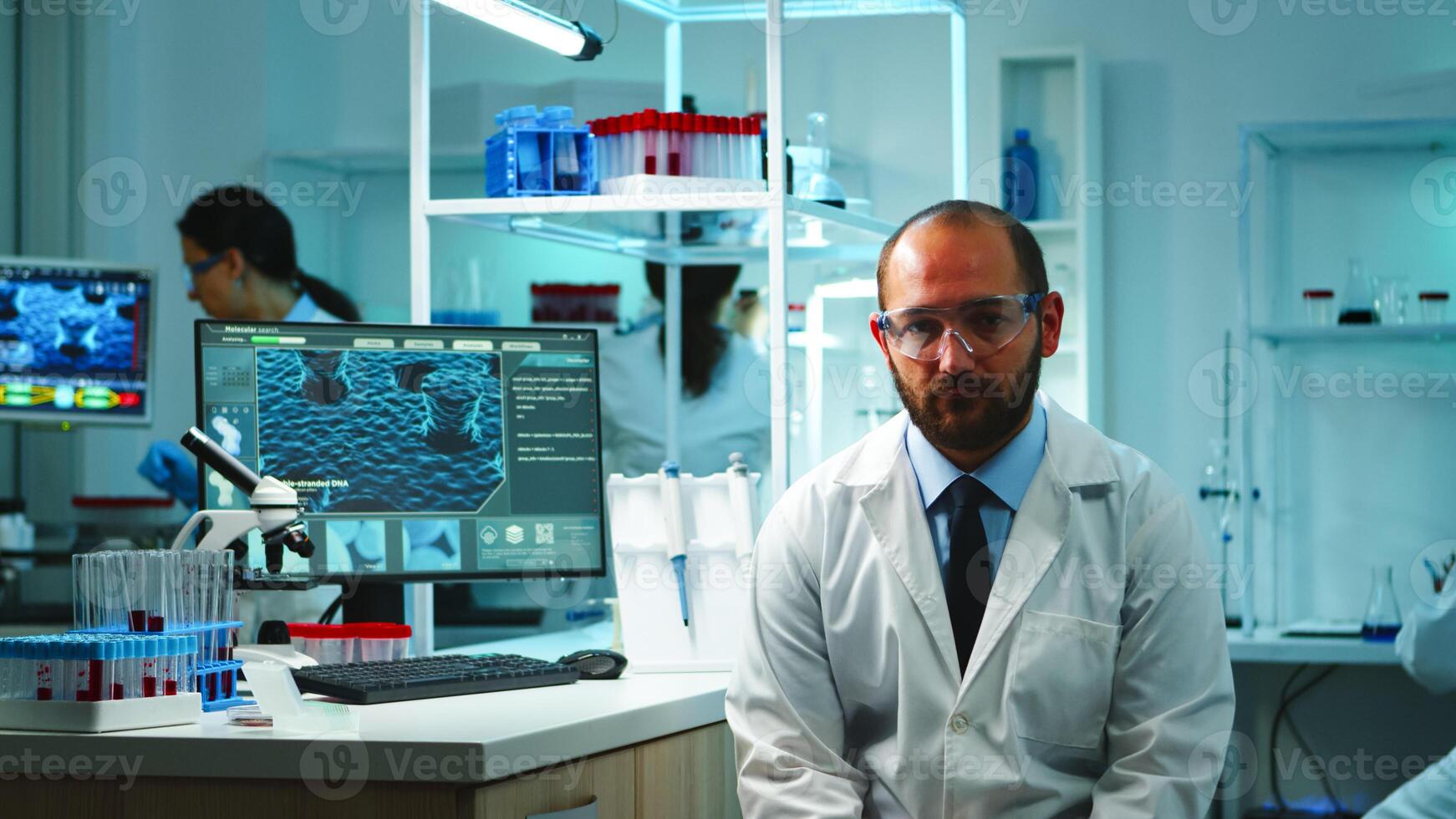 Tired doctor looking exhausted at camera in modern equipped lab late at night. Medical chemist team examining virus evolution using high tech and chemistry tools for research, vaccine development. photo
