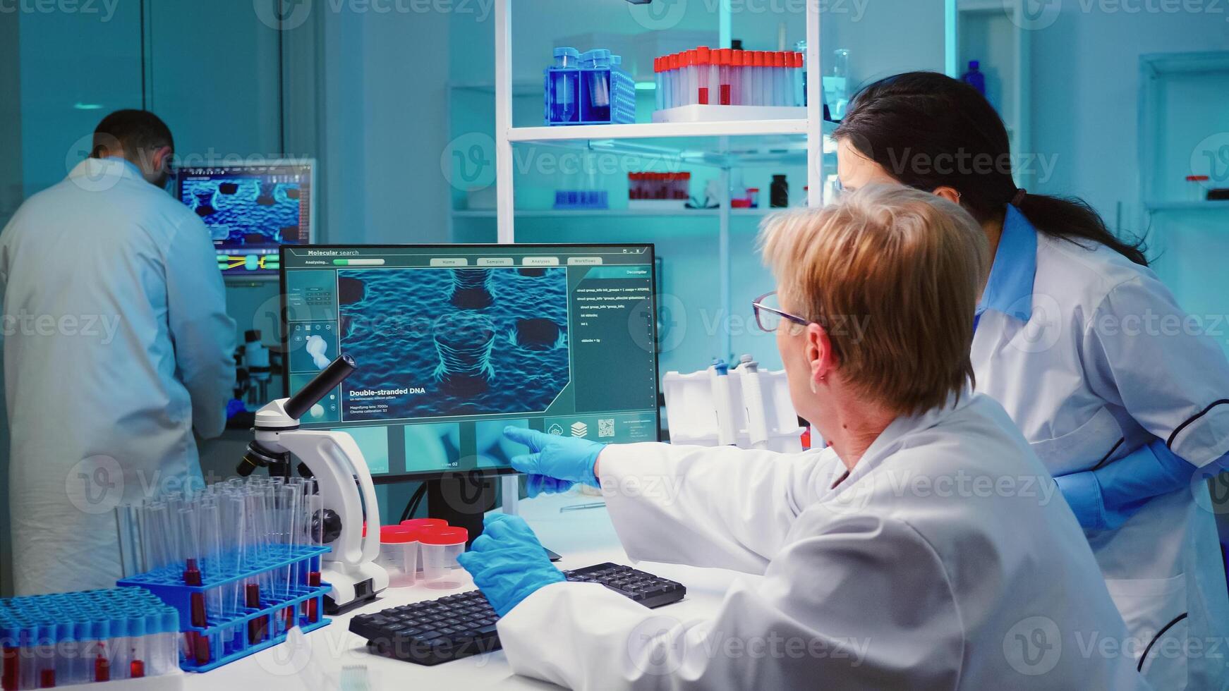 Senior biochemist writing on computer the changes in vaccine composition in modern equipped laboratory consulting with the co-worker. Examining virus evolution using high tech researching diagnosis photo