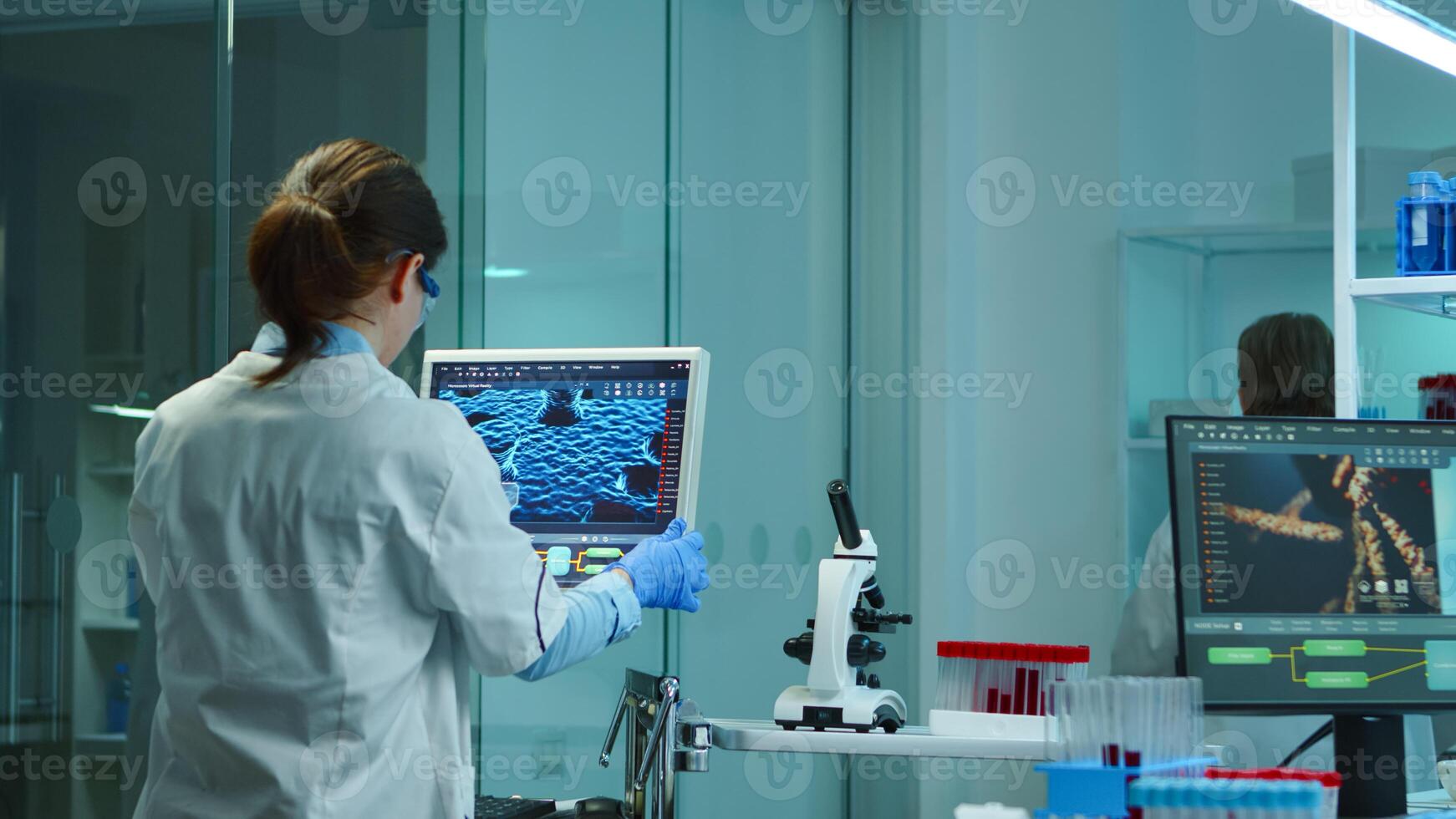 Young medical nurse positing medical monitor working late at night in modern equpped laboratory analysing virus development. Team of microbiology scientists research for vaccine against covid-19 photo