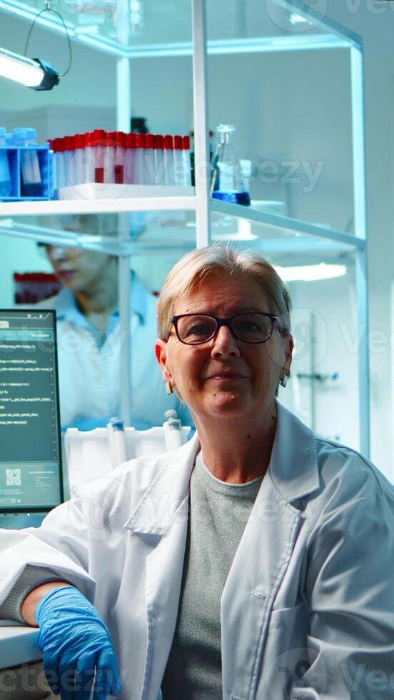 Portrait of senior scientist woman looking exhausted at camera sitting in equipped lab late at night. Team examining virus evolution using high tech for scientific research, vaccine development photo