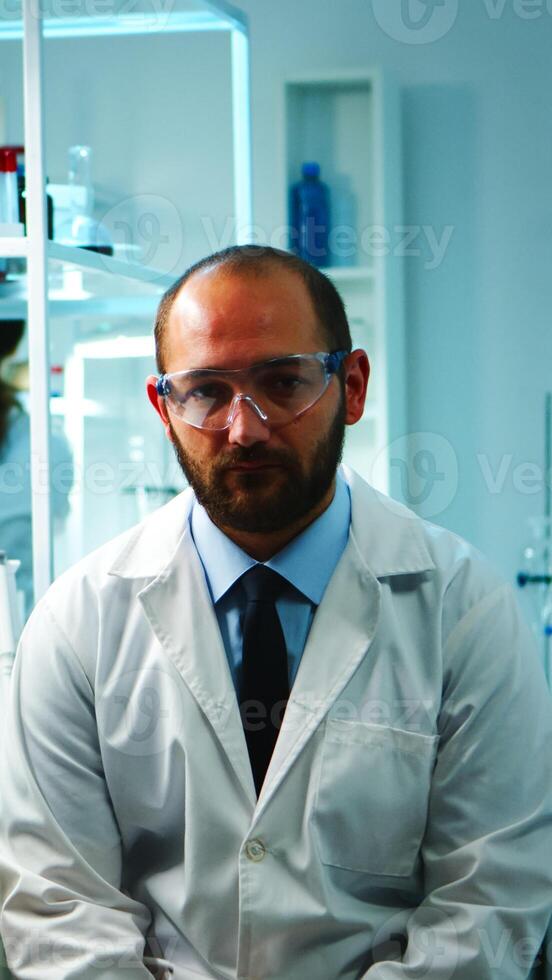 Tired doctor looking exhausted at camera in modern equipped lab late at night. Medical chemist team examining virus evolution using high tech and chemistry tools for research, vaccine development. photo