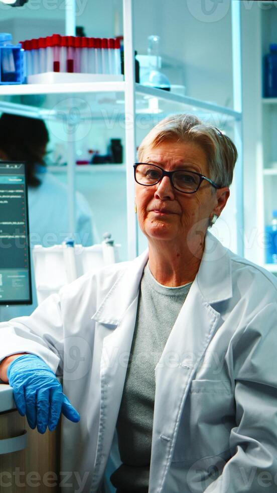 Portrait of old chemist woman with fatigued face in front of camera in equipped lab late at night. Team examining virus evolution using high tech and chemistry tools for research, vaccine development photo