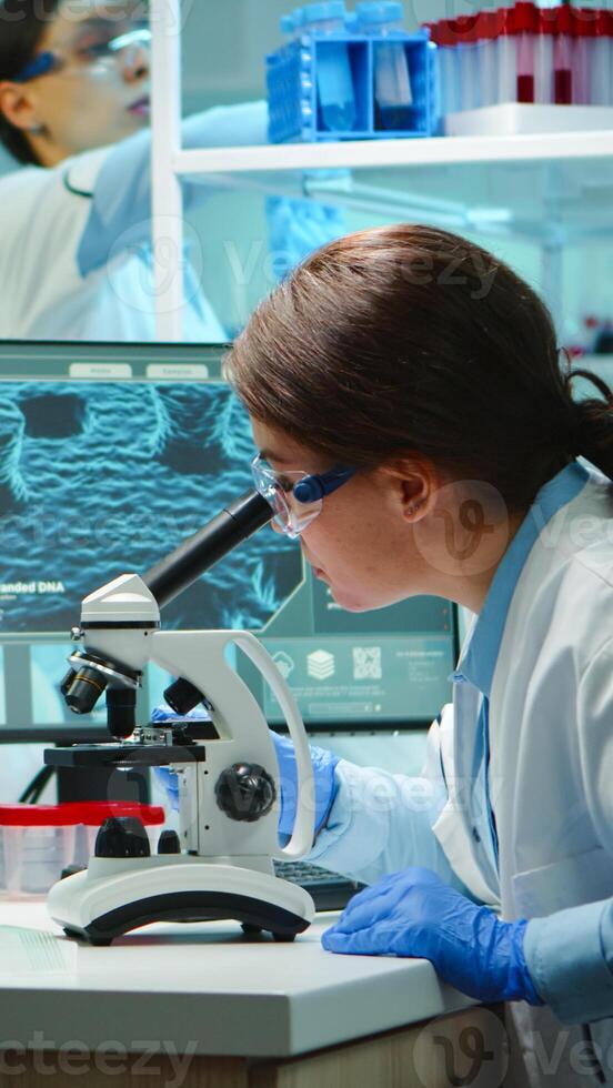 Microbiologist working late at night for new vaccine in modern laboratory looking at samples under microscope. Coworkers examining virus evolution using high tech and chemistry tools for research. photo