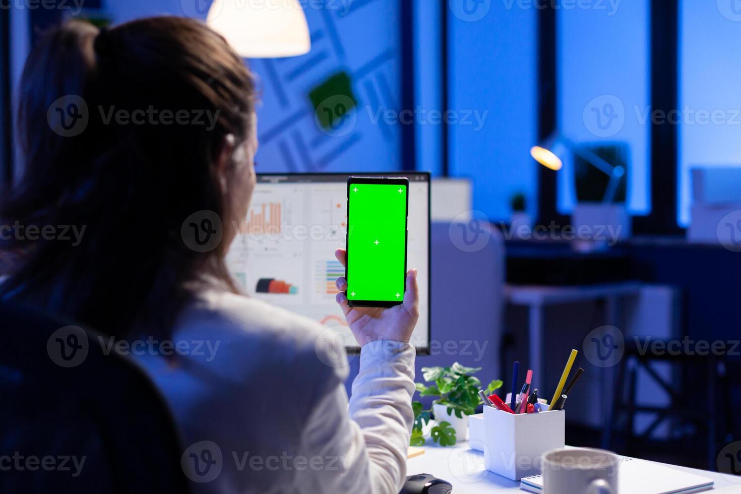 Entrepreneur looking at smartphone with green screen sitting at office desk overworking. Manager project watching desktop monitor display with mockup chroma key during videocall meeting remote photo
